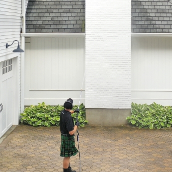 A Men In Kilts technician using a pressure washer to reach high areas on a home.